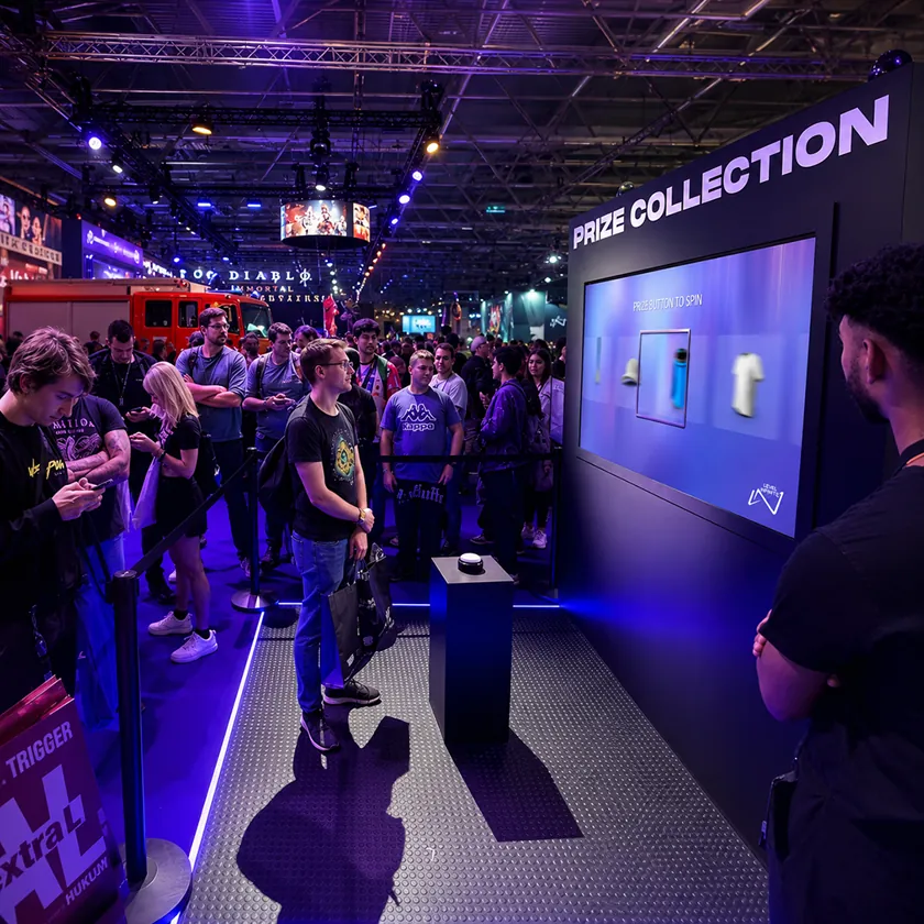 Gaming expo stand with a large prize collection screen showing a spinning merchandise reel, a big push button on a plinth, and visitors queuing behind a rope line