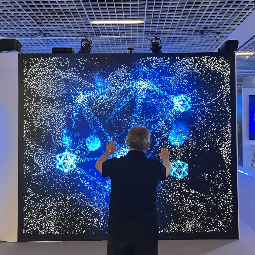 Man standing with both hands raised in front of an LED wall of white particles and glowing blue network shapes