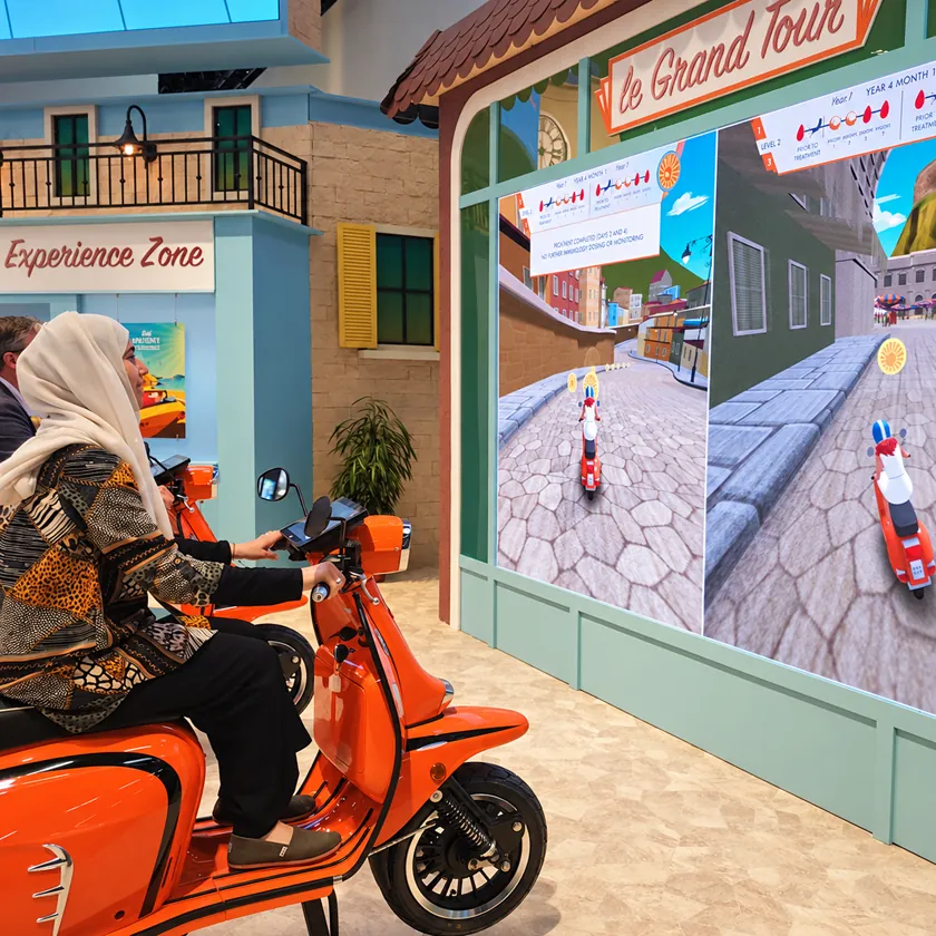 Visitor standing on a stationary scooter mounted on a platform, steering a character through a game on a large screen