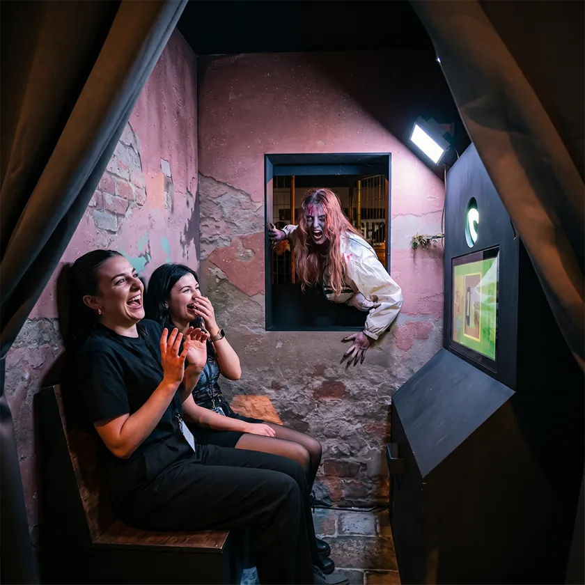 Two guests laughing and recoiling on a bench inside a dimly lit, distressed-brick booth as a zombie actor lunges through a window behind them
