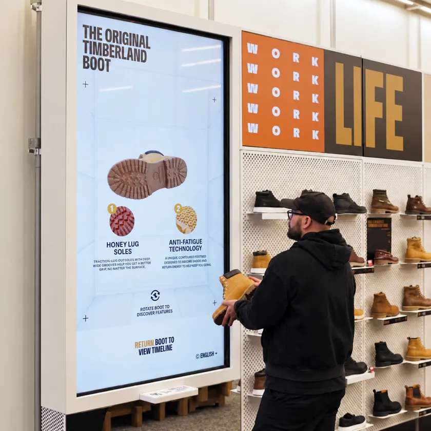 Shopper holding a shoe while a large screen beside them shows a 3D model of it turning at exactly the same angle